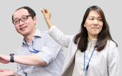 ビジネスシーンで、男性と女性が並んで立っています。女性は何かを指し示しており、男性は笑顔でカメラを見ています。