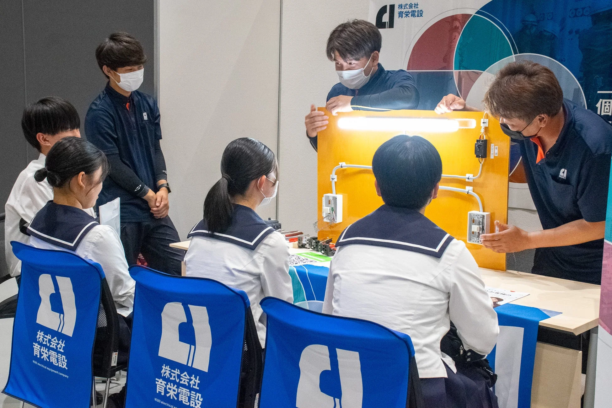 マスクを着用した育栄電設のスタッフが、学生たちに電気配線や照明の仕組みを実演で教えている様子。青い椅子には会社のロゴが描かれており、学生たちは熱心に説明を聞いている。職業体験や企業説明会のような教育的な場面を捉えた一枚。