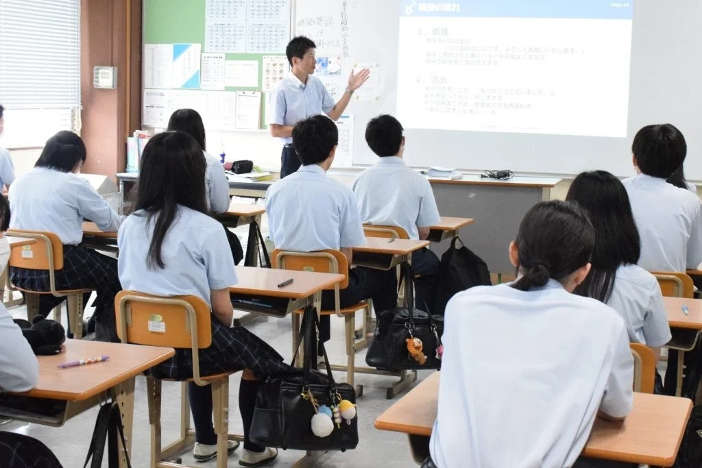 日本の学校の教室で、教師が生徒たちにプレゼンテーションを行っている様子。生徒たちは制服を着用し、熱心に授業を受けています。