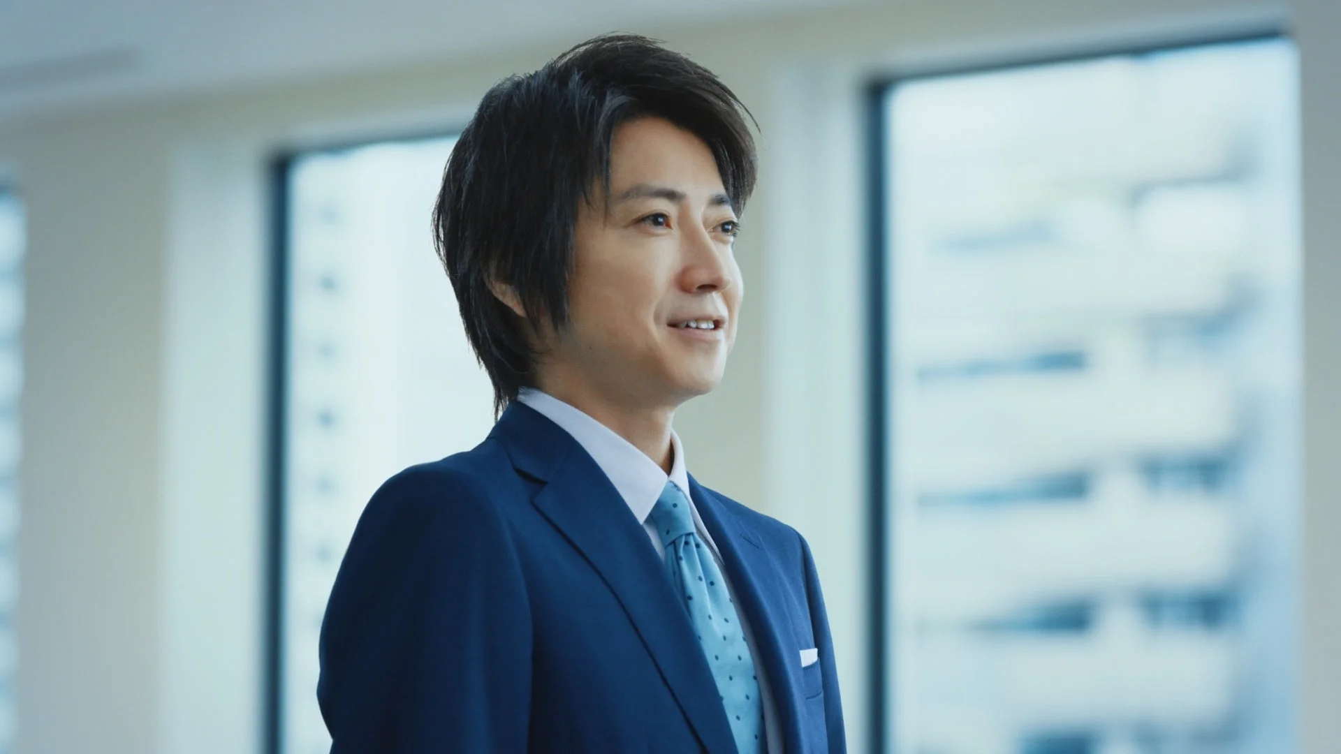 紺色のスーツを着た日本人男性が、オフィスのような場所で優しい笑顔を見せています。窓の外に視線を向けているようで、プロフェッショナルかつ信頼感のある印象を与えます。