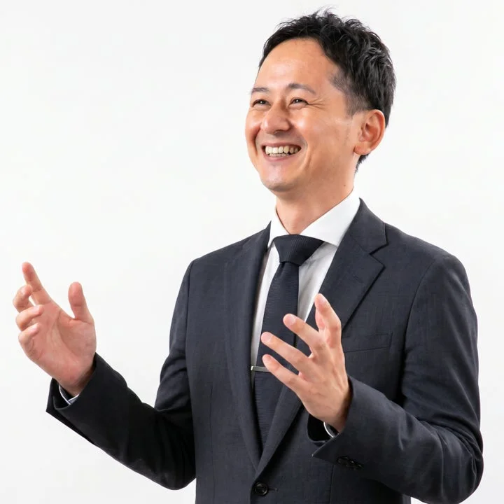 白い背景の前で、スーツ姿の日本人男性が笑顔で身振り手振りを交えながら話している様子。