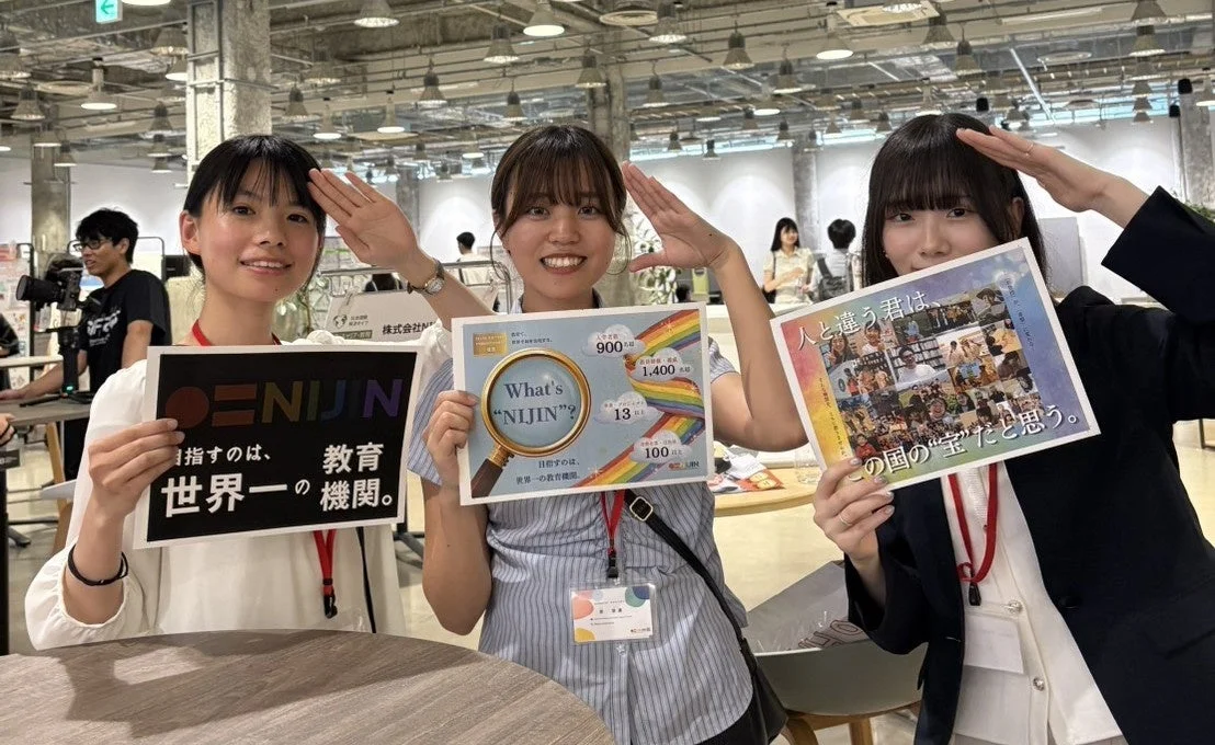 3人の若い女性が、教育機関「NIJIN」の目標や多様性を尊重するメッセージが書かれたボードを持ち、笑顔でポーズをとっています