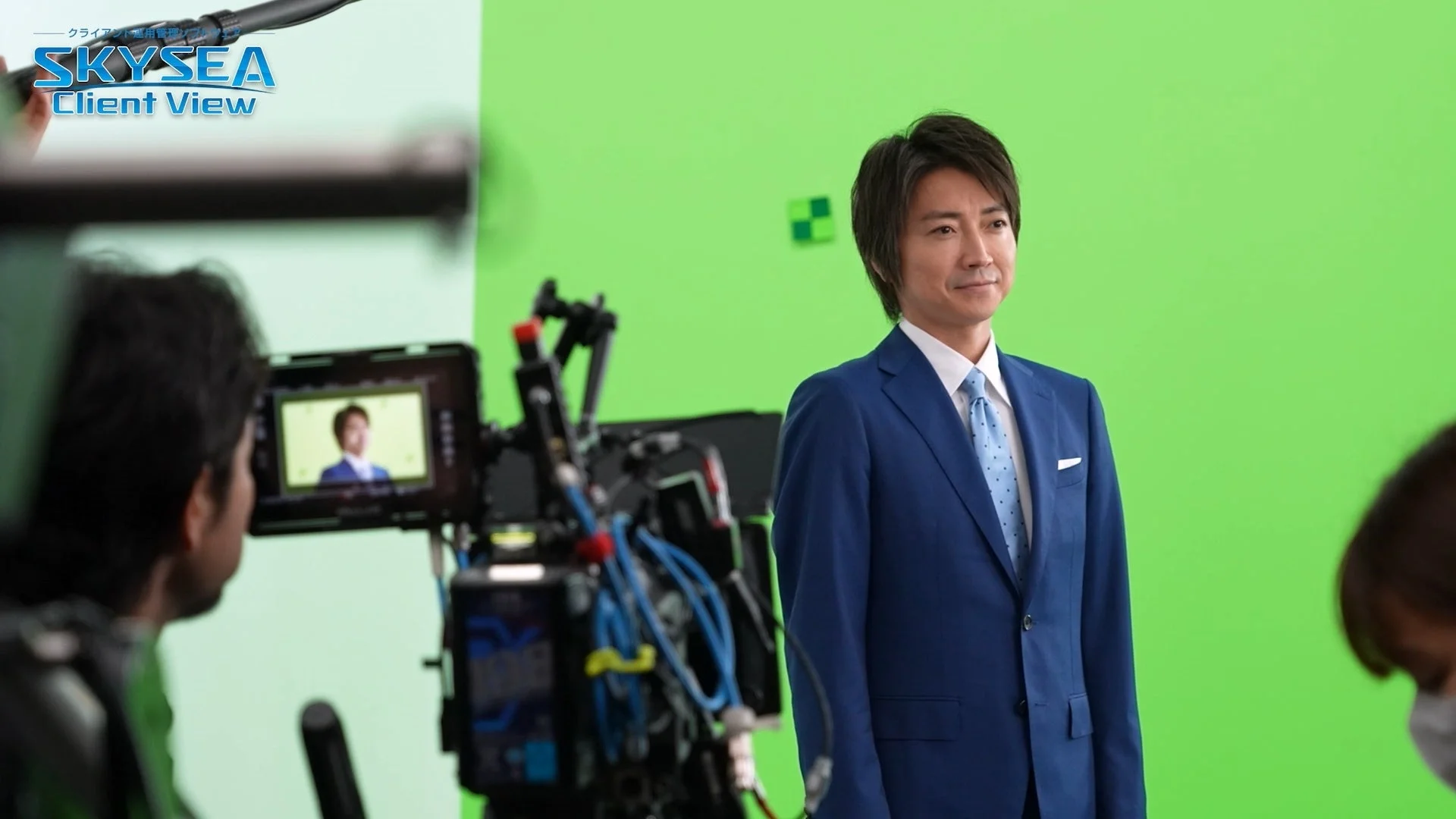 青いスーツを着た男性がグリーンバックの前で撮影されている様子です。カメラとモニターが設置されており、モニターには撮影中の男性の姿が映し出されています。