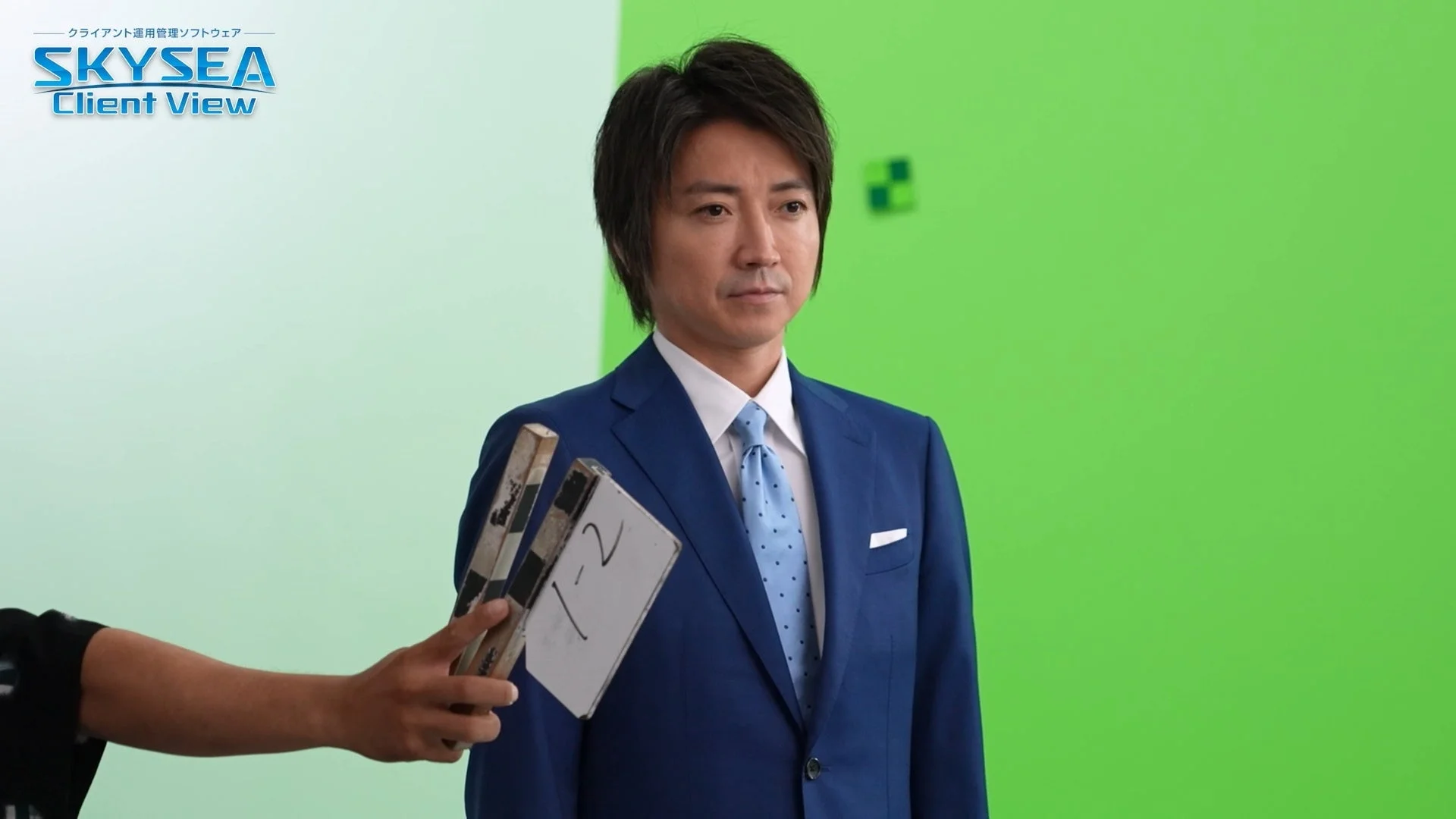 青いスーツを着た男性がグリーンスクリーンを背景に撮影されている場面。手前にはクラッパーボードがあり、左上には「SKYSEA Client View」のロゴが見えます。