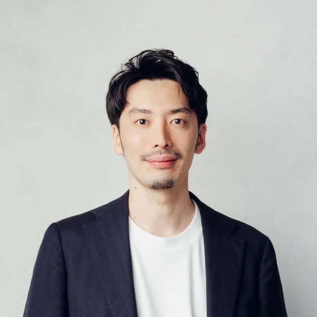 明るい背景を背に、黒いジャケットと白いTシャツを着用したアジア系の男性のポートレート