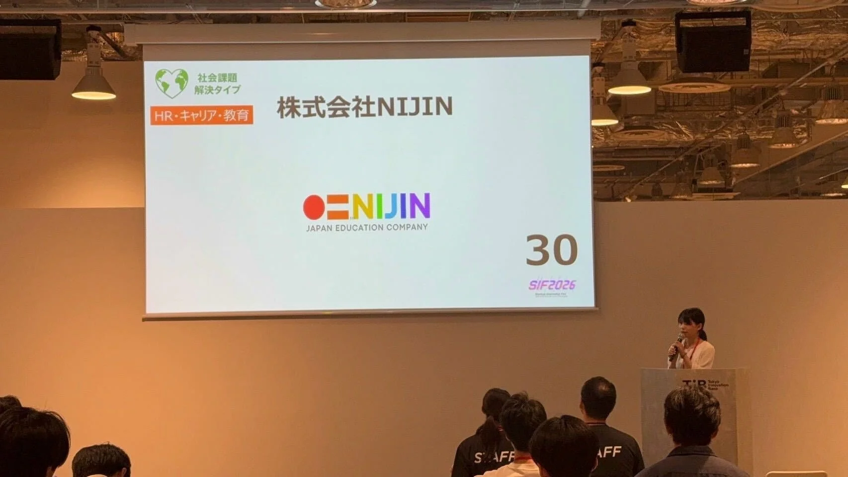 屋内で開催されているイベントで、女性がマイクを持ってプレゼンテーションを行っています