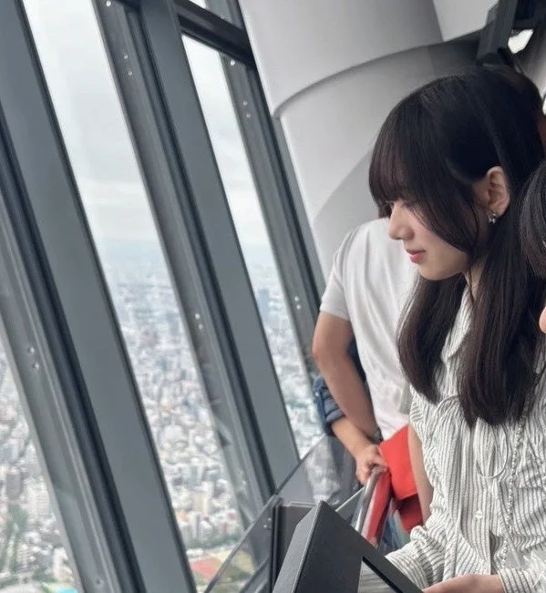 高層ビルの展望台らしき場所から、窓の外に広がる都市の景色を眺める若い女性の横顔